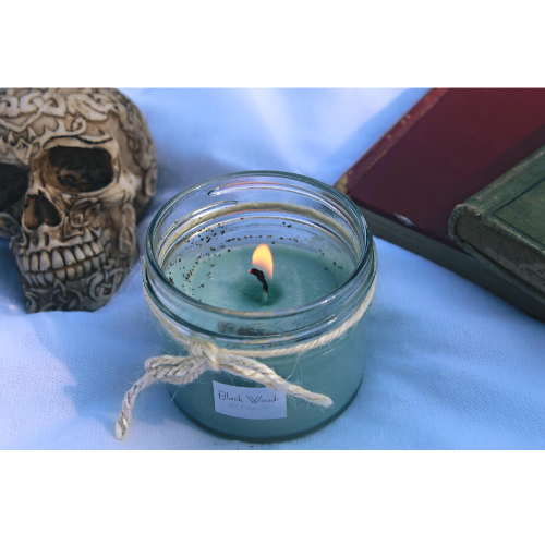 Black Woods Candles by Gravely Goods, in the background there is a skull and old books, everything is on top of cloth
