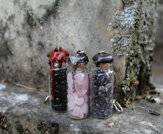 Obsidian, rose quartz, and amethyst crystals inside clear glass jar with wax seals and keychain attachments in front of an old tombstone