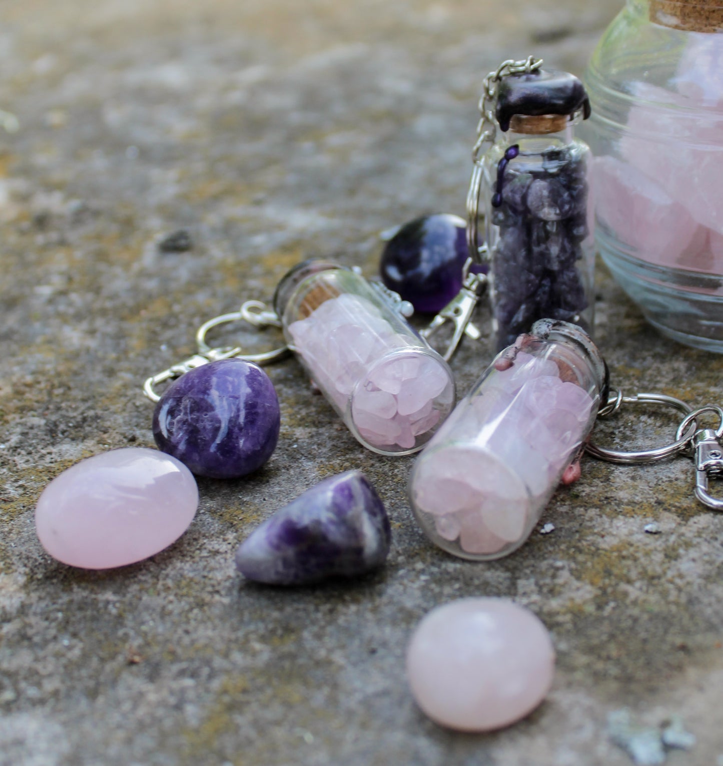 rose quartz, and amethyst crystals inside clear glass jar with wax seals and keychain attachments in front of an old tombstone by gravely goods scattered next to rose quartz, and amethyst tumbled stones